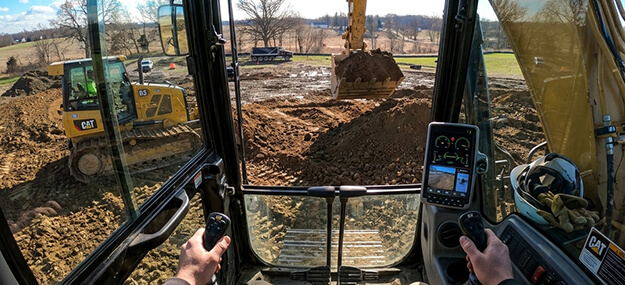 View from inside excavator cab showing operator controls and active site preparation work with heavy equipment moving soil