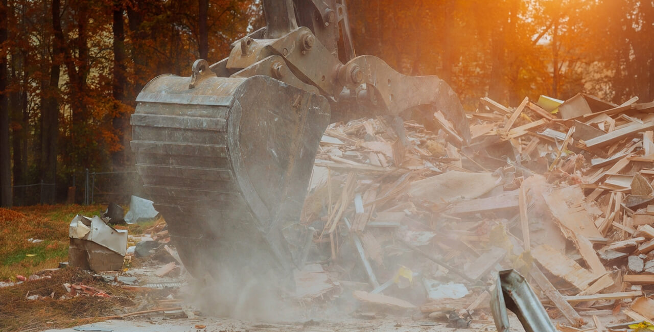 Excavator breaking down debris during residential demolition with dust and material removal
