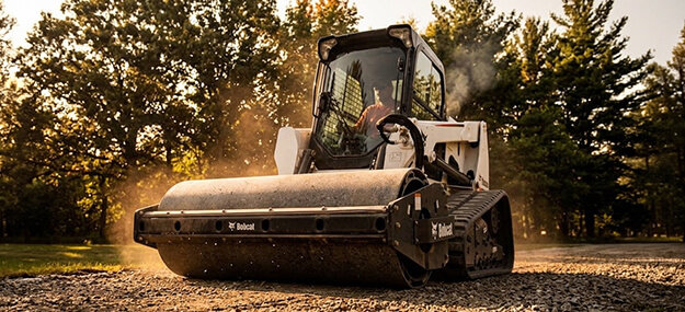 Skid steer with roller attachment compacting gravel base during site preparation for concrete flatwork