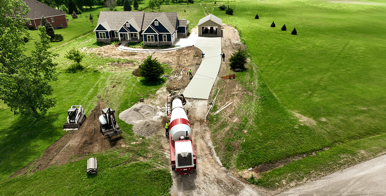 Aerial view of residential concrete driveway installation with cement truck, excavator, and crew working on a new home site