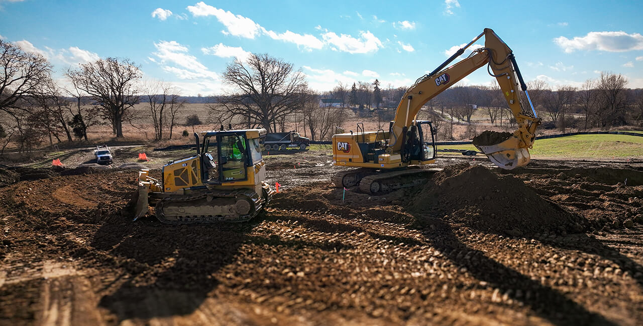 Excavator and bulldozer performing site preparation and grading work on a rural construction site with open land and trees